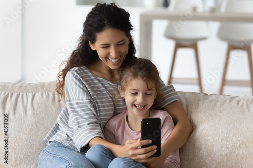Overjoyed young Latino mother and daughter use cellphone look at screen laugh watch funny video online. Smiling Hispanic mom and small girl child talk speak on webcam virtual event on smartphone.