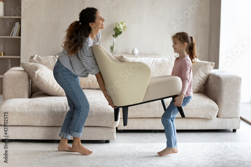 Smiling young single Hispanic mother and teen daughter carry armchair settle in new own home. Happy Latino mom and teenage girl child decorate cozy living room. Moving, relocation concept.