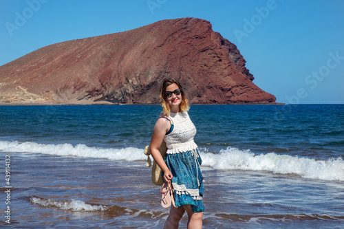 Portrait of an exuberant young millennial woman wearing a blue and white paisley dress while smiling, walking and enjoying her summer vacation days at La Tejita beach, Tenerife, Canary Islands, Spain photo