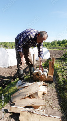  A 60-year-old Caucasian man in working clothes, shod in black boots, shows how to gradually form firewood from birch using an old device, tying them with a natural rope. 