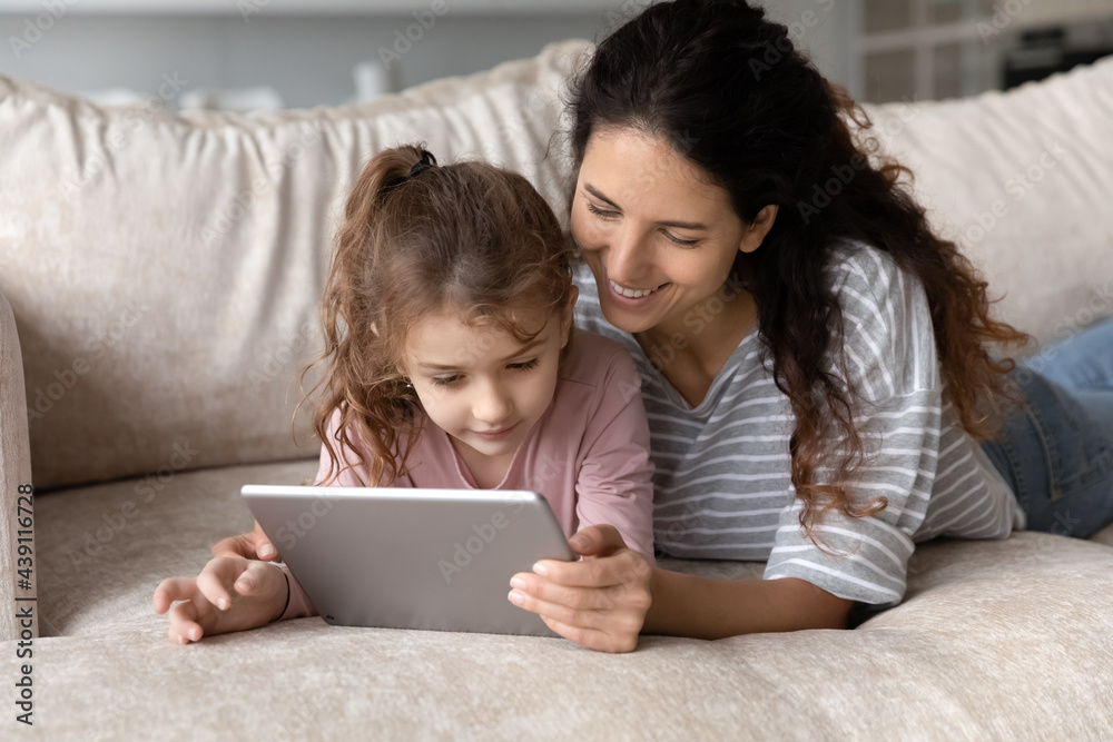 Happy loving young Latino mother and daughter lying relaxing on sofa at use modern tablet gadget together. Smiling Hispanic mom and small girl child look at pad screen browse internet on device. Happy loving young Latino mother and daughter lying relaxing on sofa at use modern tablet gadget together. Smiling Hispanic mom and small girl child look at pad screen browse internet on device.