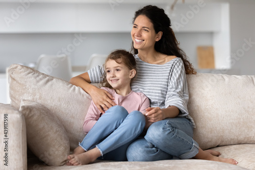 Smiling Hispanic young mother and happy small biracial daughter sit on sofa at home look in distance dream visualize together. Excited Latino mom and little ethnic girl child relax. Vision concept.