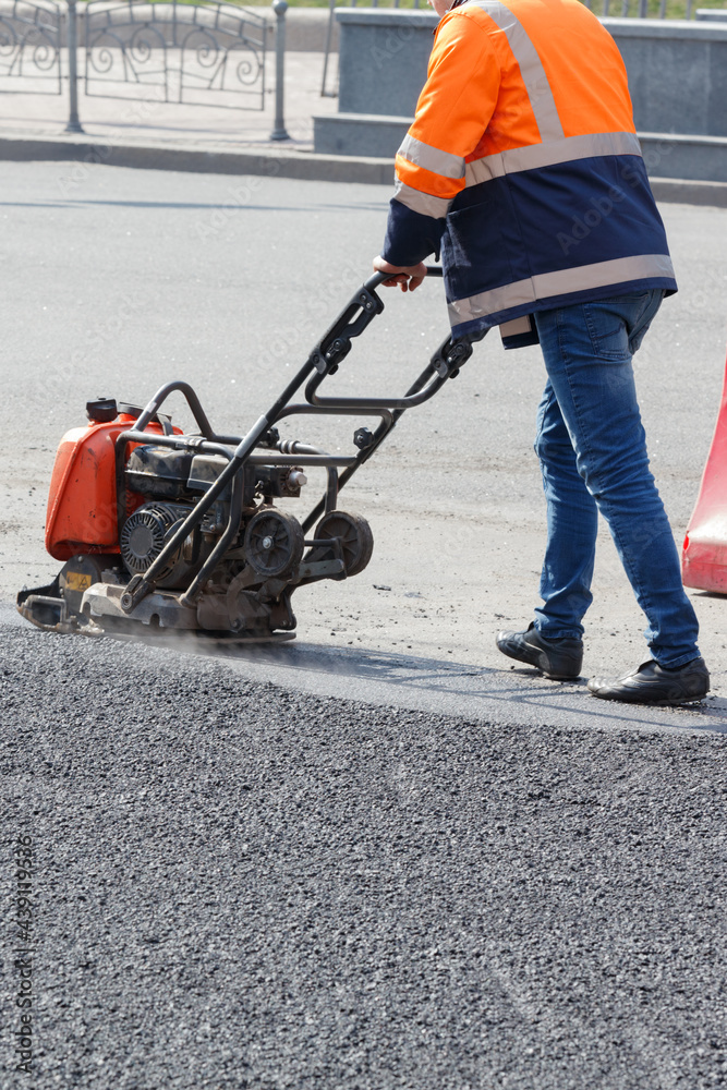 Fototapeta premium A road worker compacts fresh asphalt with petrol vibratory plate on a sunny day.