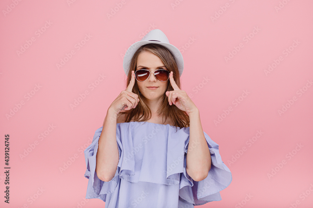 Cheerful female in stylish top with bare shoulders going to have walk at beach wears sunglasses looking at camerastanding isolated against a pink background