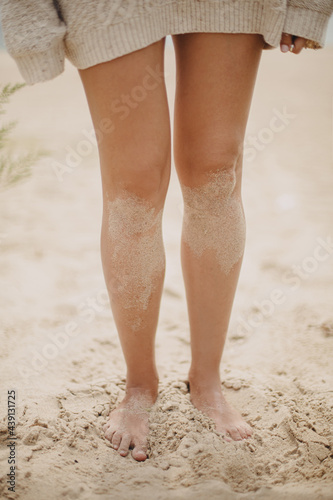 Beautiful woman legs barefoot with sand standing on sandy beach, carefree vacation mood. Stylish young female in knitted sweater relaxing on coast, cropped view. Vertical image