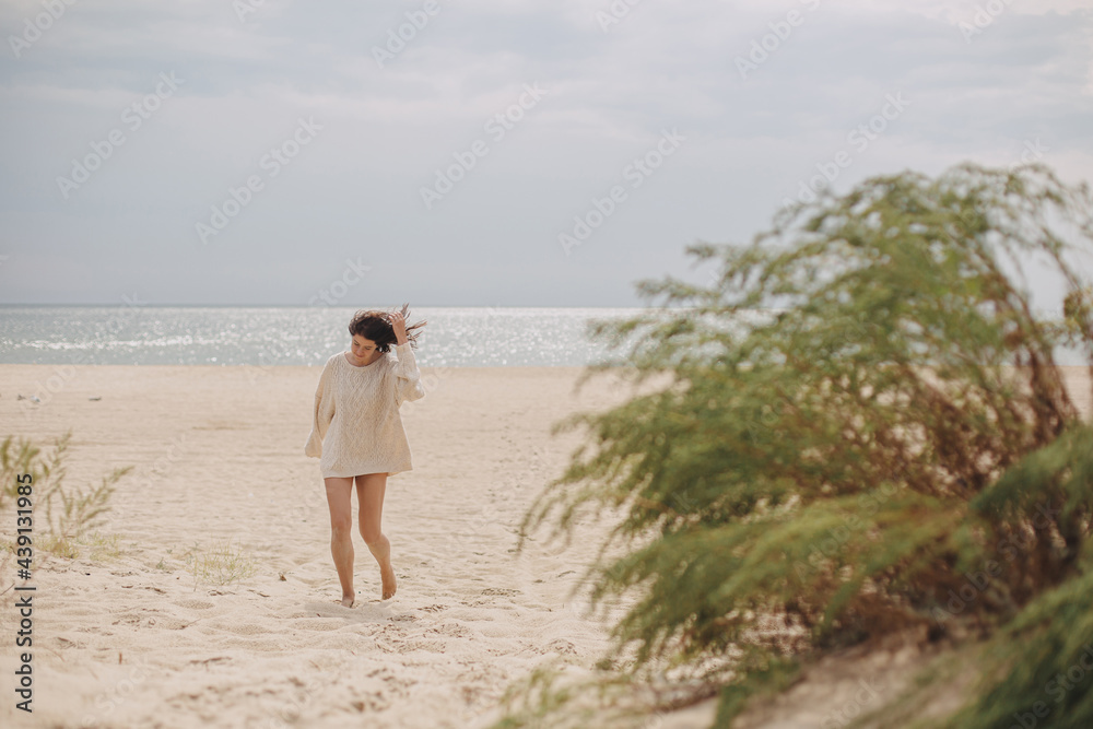 Beautiful woman with windy hair walking on sandy beach on background of green grass and sea, calm tranquil moment. Stylish young female in beige knitted sweater relaxing on cold coast