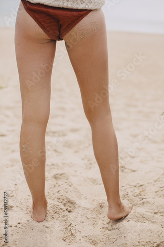 Woman buttocks and tanned legs in sand on background of sandy beach, lifestyle. Stylish young female in sweater and swimsuit with sandy buttocks relaxing on coast. Summer beach. Vertical image