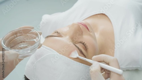 Overhead view of young woman relaxing on massage table during beauty spa procedure and doctor cosmetologist applying moisturizing cream on her face. Concept of care for face skin. 