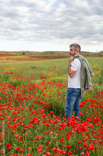 A man in a white t-shirt and jeans stands in a blooming poppy field and looks into the distance. 
Summer countryside vacation theme.