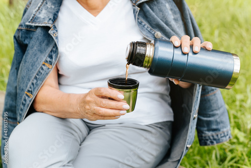 Outdoor camping. Senior woman having a rest in nature. Elderly woman's hands holding a thermos and pouring tea into a mug. Cropped image