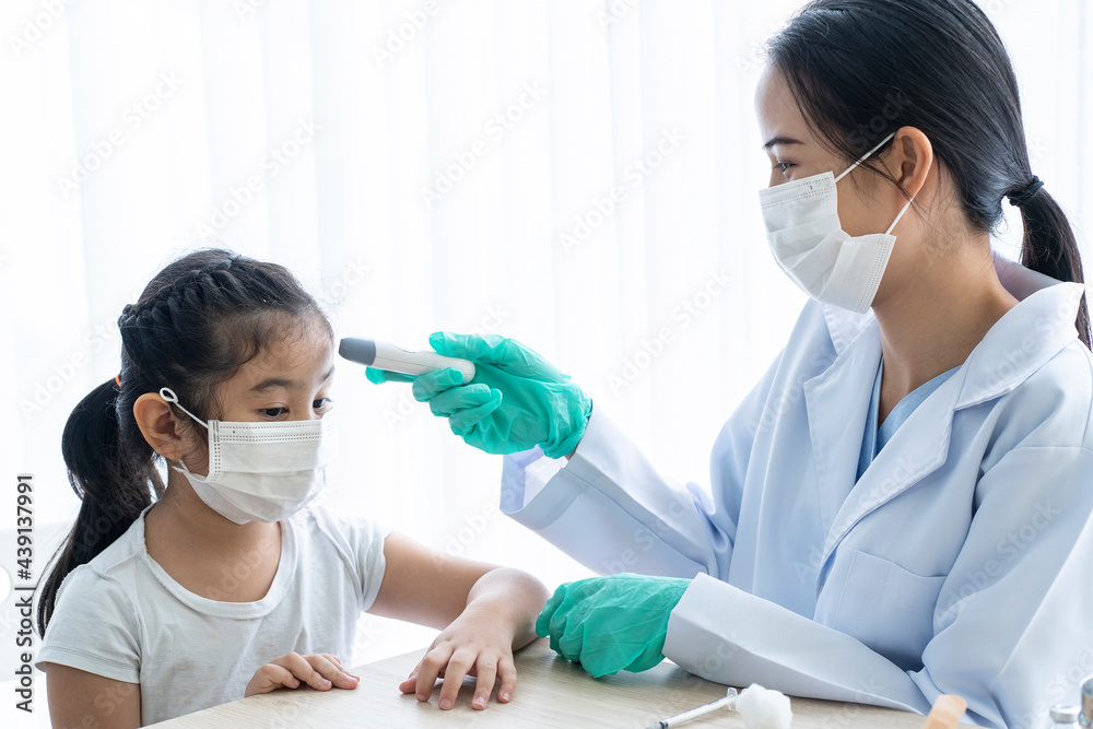 An Asian female doctor sits and examines a young girl's temperature in a clinic.