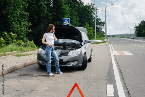 A young girl stands near a broken-down car in the middle of the highway and calls for help on the phone. Failure and breakdown of the car. Waiting for help. © Andrii