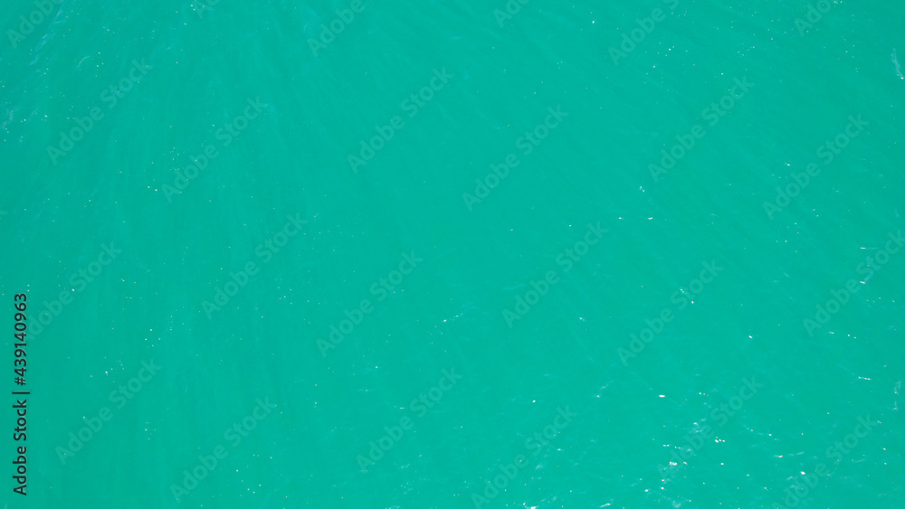 Top view of green water and small waves. The background of the water shimmers in the rays of the sun with highlights. White foam is visible. The gradient of the lake and the beach. Rocks on the bottom Top view of green water and small waves. The background of the water shimmers in the rays of the sun with highlights. White foam is visible. The gradient of the lake and the beach. Rocks on the bottom