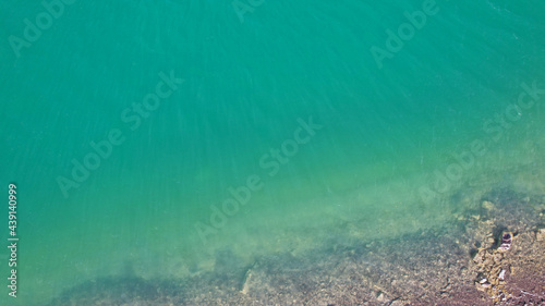 Top view of green water and small waves. The background of the water shimmers in the rays of the sun with highlights. White foam is visible. The gradient of the lake and the beach. Rocks on the bottom