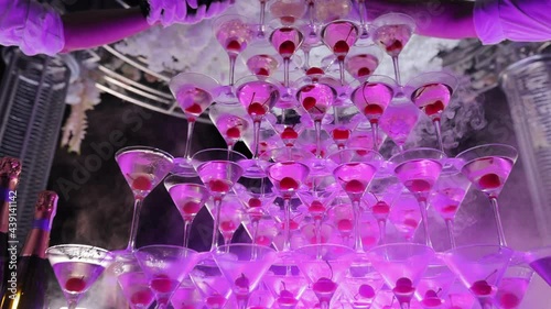 Waiter pours champagne in glass tower. Tower of champagne glasses with two bottles on the table. Pyramid of glasses for new year eves party. Restaurant service of champagne tower at the background