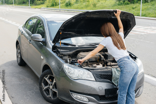 A young girl stands near a broken-down car in the middle of the highway and looks under the hood. Failure and breakdown of the car. Waiting for help. © Andrii