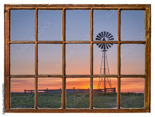 windmill with a water pump and tank against sunrise in a prairie as seen from vintage sash window