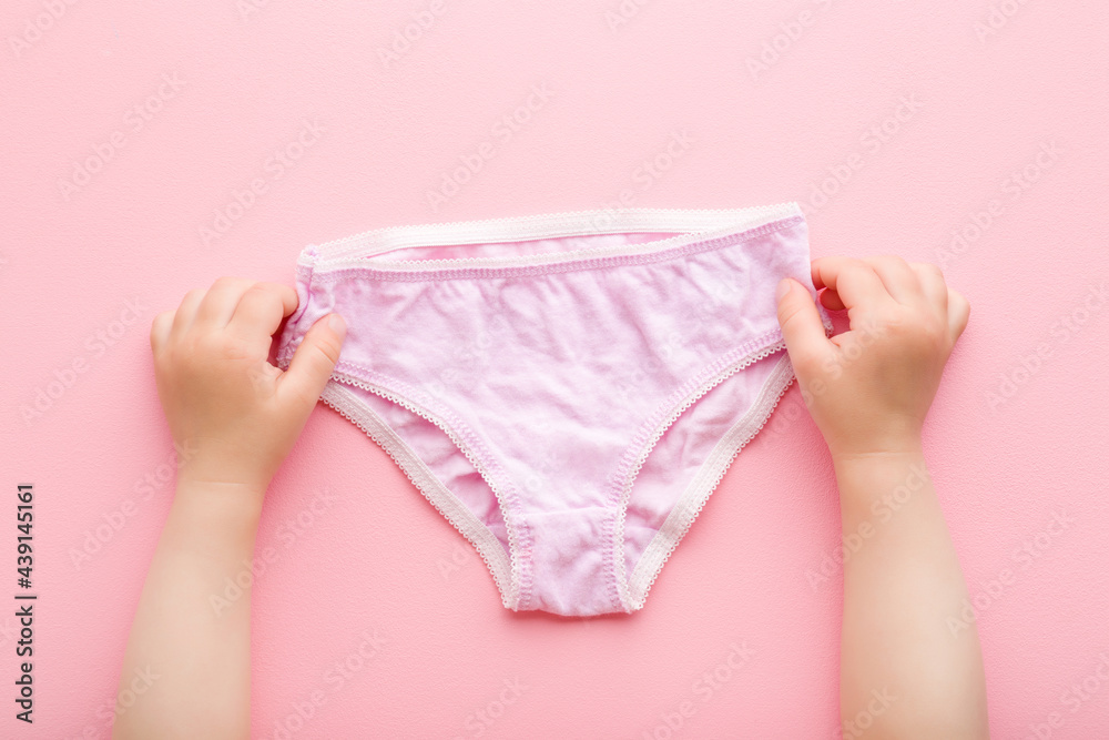 Baby girl hands holding on light purple panties on pink table background. Pastel color. Closeup. Point of view shot. First underwear after diaper using. Top down view. - obrazy, fototapety, plakaty 