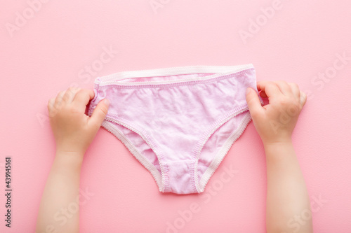 Baby girl hands holding on light purple panties on pink table background. Pastel color. Closeup. Point of view shot. First underwear after diaper using. Top down view.