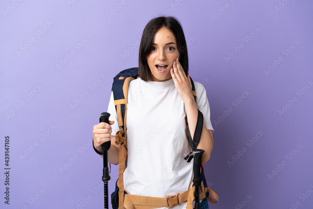 Young caucasian woman with backpack and trekking poles isolated on blue background with surprise and shocked facial expression