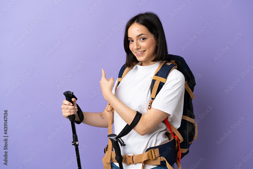 Young caucasian woman with backpack and trekking poles isolated on blue background pointing back