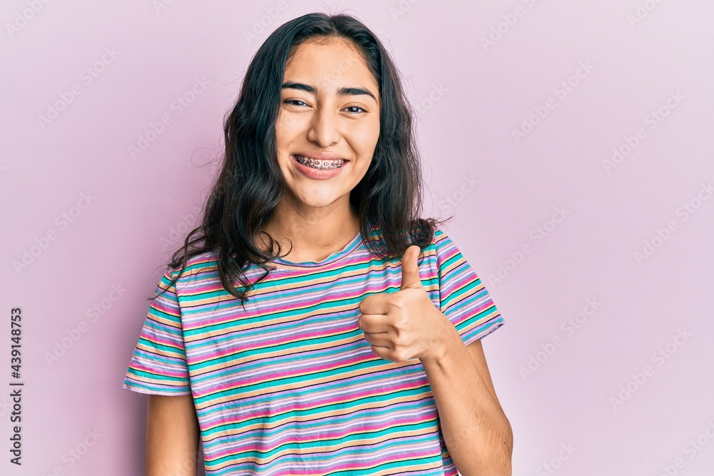 Hispanic teenager girl with dental braces wearing casual clothes doing happy thumbs up gesture with hand. approving expression looking at the camera showing success. Hispanic teenager girl with dental braces wearing casual clothes doing happy thumbs up gesture with hand. approving expression looking at the camera showing success.