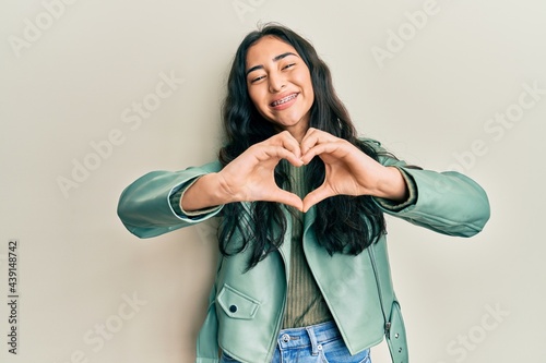 Hispanic teenager girl with dental braces wearing green leather jacket smiling in love doing heart symbol shape with hands. romantic concept.