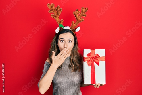 Young hispanic girl wearing deer christmas hat holding gift covering mouth with hand, shocked and afraid for mistake. surprised expression