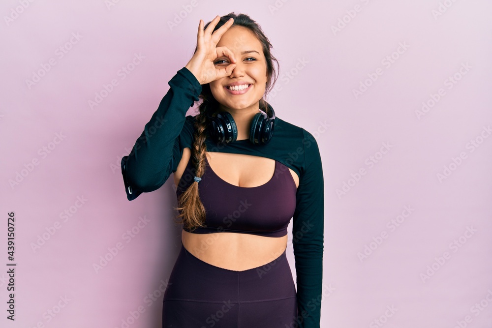 Obraz premium Young hispanic girl wearing sportswear and headphones doing ok gesture with hand smiling, eye looking through fingers with happy face.