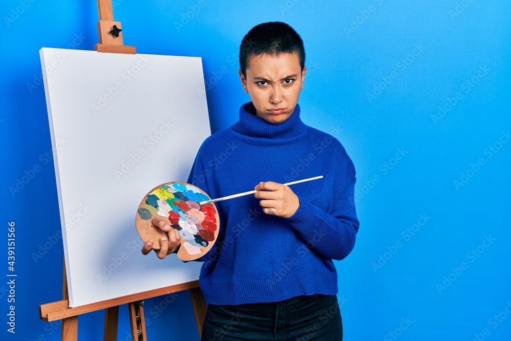 Fototapeta premium Beautiful hispanic woman with short hair holding paintbrush and palette skeptic and nervous, frowning upset because of problem. negative person.