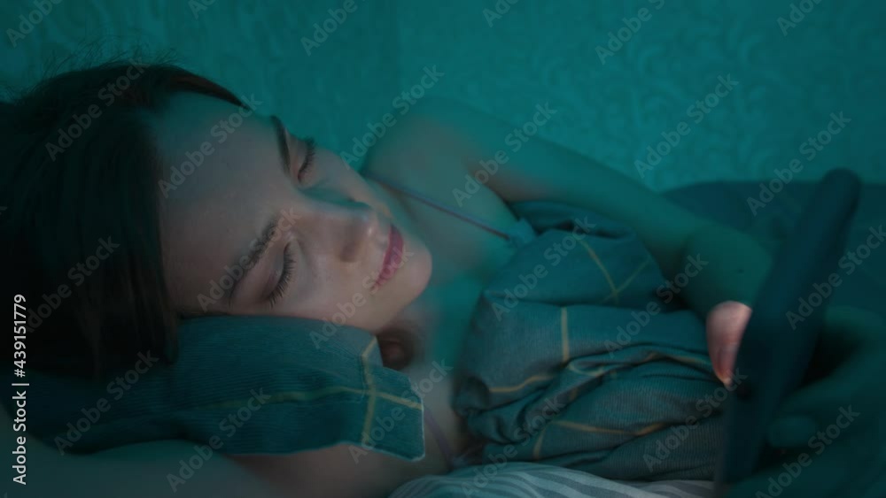 Young caucasian woman lying in a bed in the bedroom browsing the Internet using a smartphone, sleepy tired late at night. Dramatic cold dark light. On the Internet, mobile addiction and insomnia. Young caucasian woman lying in a bed in the bedroom browsing the Internet using a smartphone, sleepy tired late at night. Dramatic cold dark light. On the Internet, mobile addiction and insomnia.