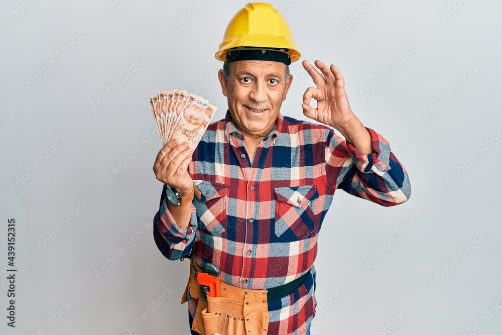 Senior hispanic man wearing handyman uniform holding turkish liras doing ok sign with fingers, smiling friendly gesturing excellent symbol