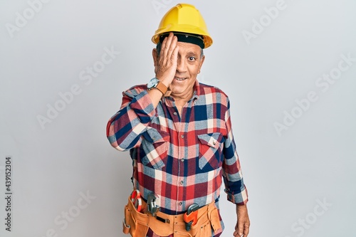 Senior hispanic man wearing handyman uniform covering one eye with hand, confident smile on face and surprise emotion.
