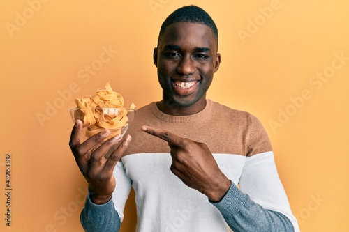 Young african american man holding bowl with uncooked spaghetti smiling happy pointing with hand and finger
