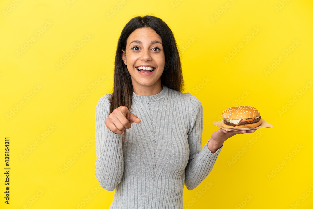 Young caucasian woman holding a burger isolated on yellow background surprised and pointing front Young caucasian woman holding a burger isolated on yellow background surprised and pointing front