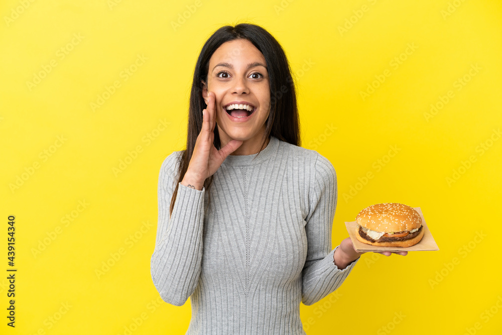 Young caucasian woman holding a burger isolated on yellow background with surprise and shocked facial expression Young caucasian woman holding a burger isolated on yellow background with surprise and shocked facial expression