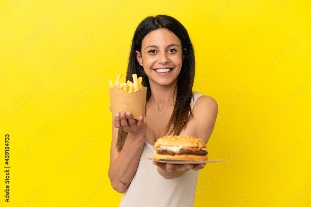 Young caucasian woman holding burger and chips isolated on yellow background Young caucasian woman holding burger and chips isolated on yellow background