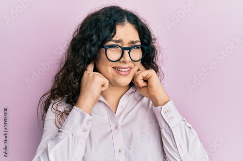 Young brunette woman with curly hair wearing casual clothes and glasses covering ears with fingers with annoyed expression for the noise of loud music. deaf concept.