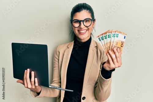 Young hispanic woman wearing business style holding laptop and 50 euros smiling with a happy and cool smile on face. showing teeth. photo