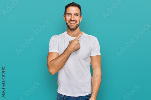 Handsome man with beard wearing casual white t shirt cheerful with a smile of face pointing with hand and finger up to the side with happy and natural expression on face
