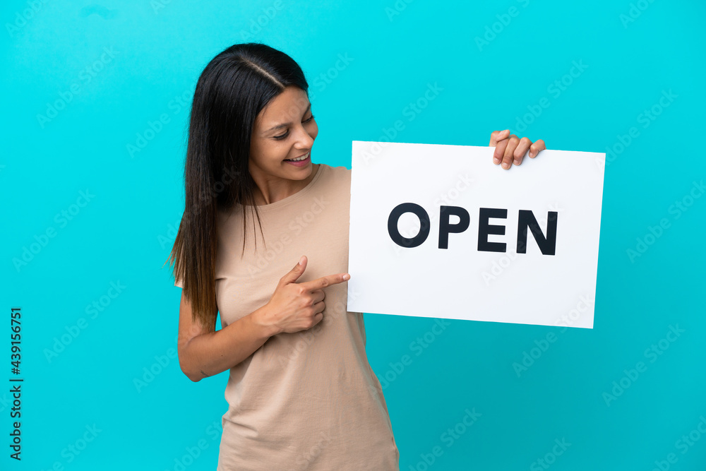 Young woman over isolated background holding a placard with text OPEN and pointing it Young woman over isolated background holding a placard with text OPEN and pointing it