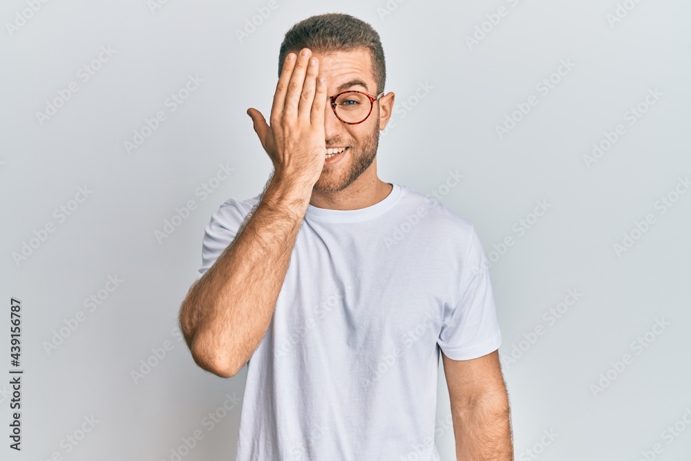 Young caucasian man wearing casual clothes and glasses covering one eye with hand, confident smile on face and surprise emotion. Young caucasian man wearing casual clothes and glasses covering one eye with hand, confident smile on face and surprise emotion.