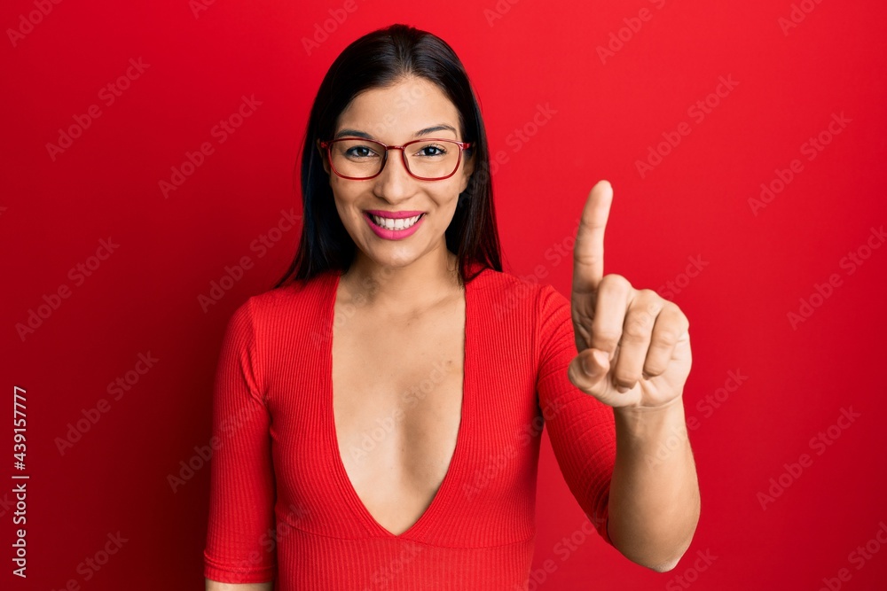 Young latin woman wearing casual clothes and glasses showing and pointing up with finger number one while smiling confident and happy. Young latin woman wearing casual clothes and glasses showing and pointing up with finger number one while smiling confident and happy.
