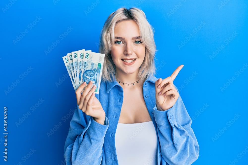 Young blonde girl holding 50 polish zloty banknotes smiling happy pointing with hand and finger to the side Young blonde girl holding 50 polish zloty banknotes smiling happy pointing with hand and finger to the side