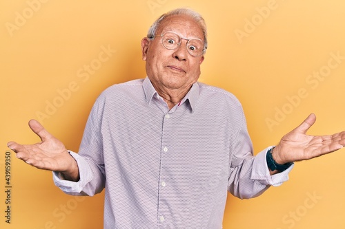 Senior man with grey hair wearing elegant shirt and glasses clueless and confused expression with arms and hands raised. doubt concept.