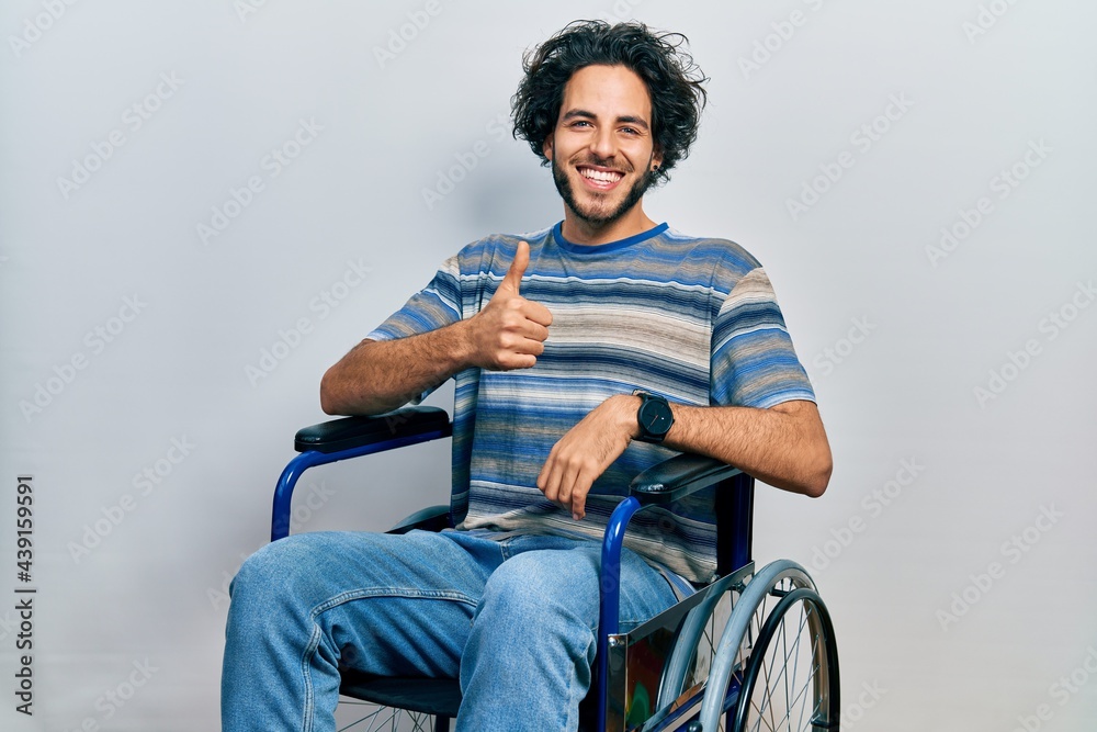 Handsome hispanic man sitting on wheelchair doing happy thumbs up gesture with hand. approving expression looking at the camera showing success.