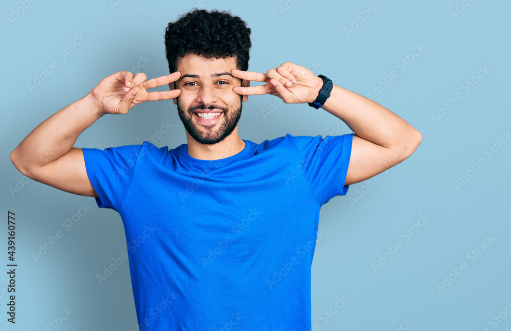 Young arab man with beard wearing casual blue t shirt doing peace symbol with fingers over face, smiling cheerful showing victory Young arab man with beard wearing casual blue t shirt doing peace symbol with fingers over face, smiling cheerful showing victory