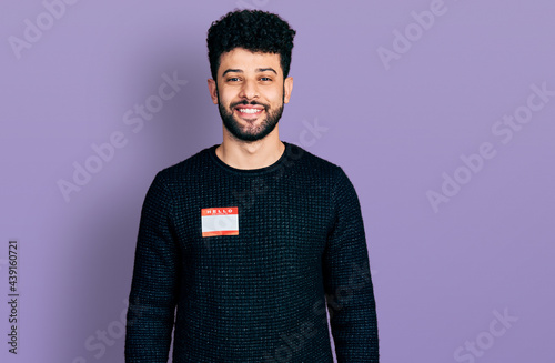 Young arab man with beard wearing hello my name is sticker identification looking positive and happy standing and smiling with a confident smile showing teeth photo