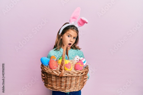 Little beautiful girl wearing cute easter bunny ears holding wicker basket with colored eggs in shock face, looking skeptical and sarcastic, surprised with open mouth