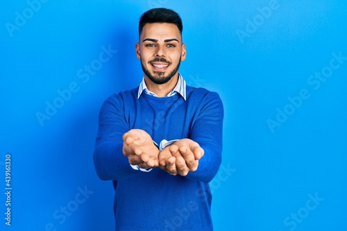 Young hispanic man with beard wearing casual blue sweater smiling with hands palms together receiving or giving gesture. hold and protection